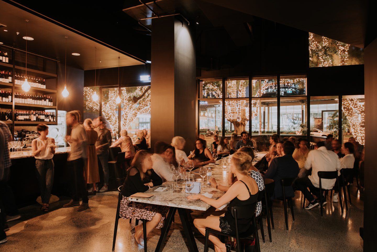 The main dining room at Snack Man, long marble tables filled with guests and fairy-lit trees glowing through the windows onto East Street