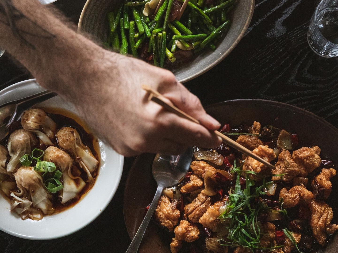 Guests sharing a banquet at Snack Man, chopsticks reaching across a dark table for stir-fried green beans, wontons in chilli oil and crispy chilli chicken